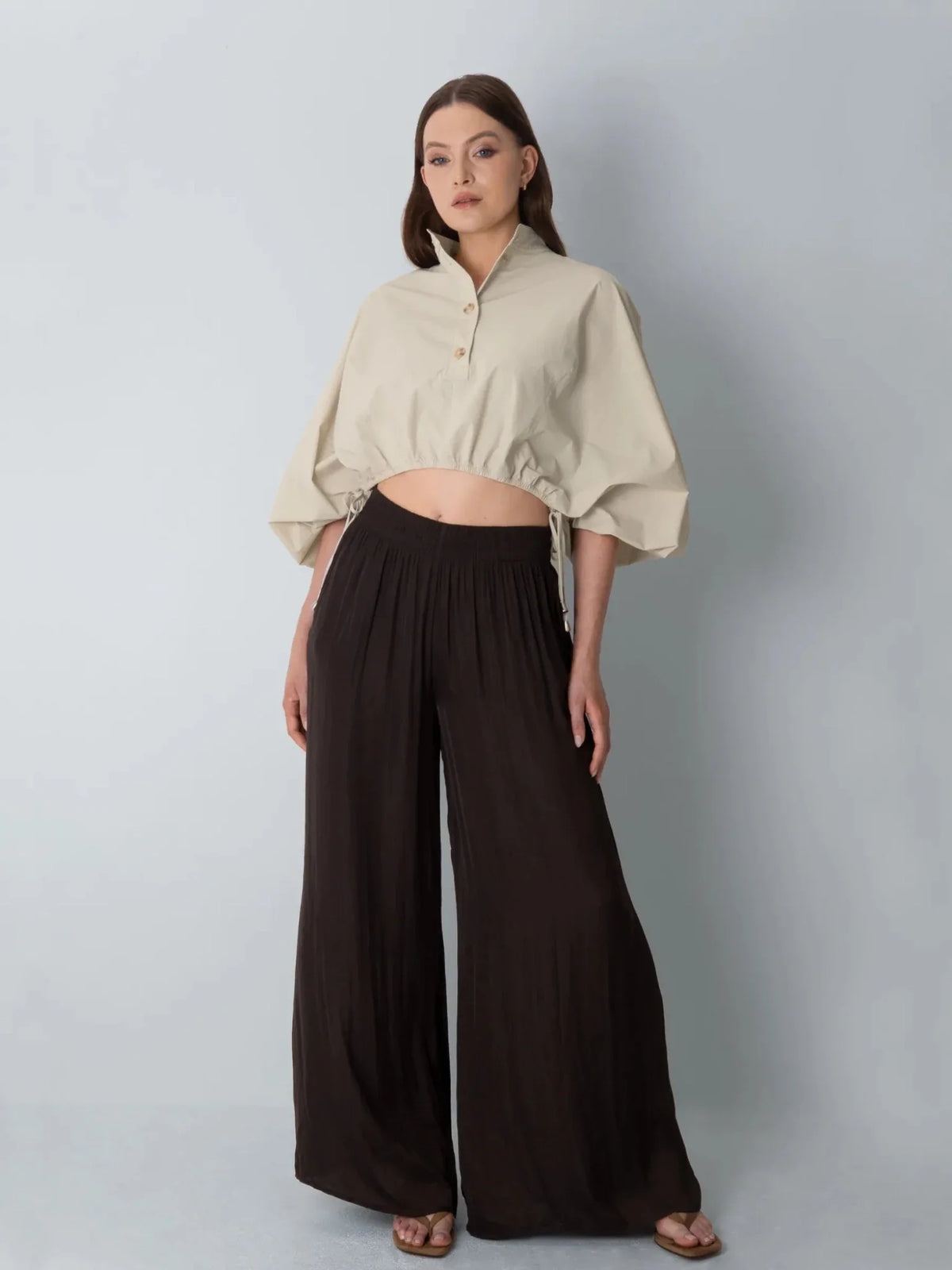 Woman wearing a PUFFLT cream drawstring waist blouse and dark brown wide-leg trousers.